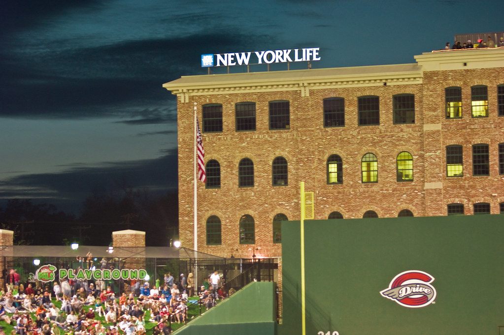 Fans on the outfield lawn at Fluor Field at dusk during a Greenville Drive baseball game, with a lit brick mill building behind the green wall