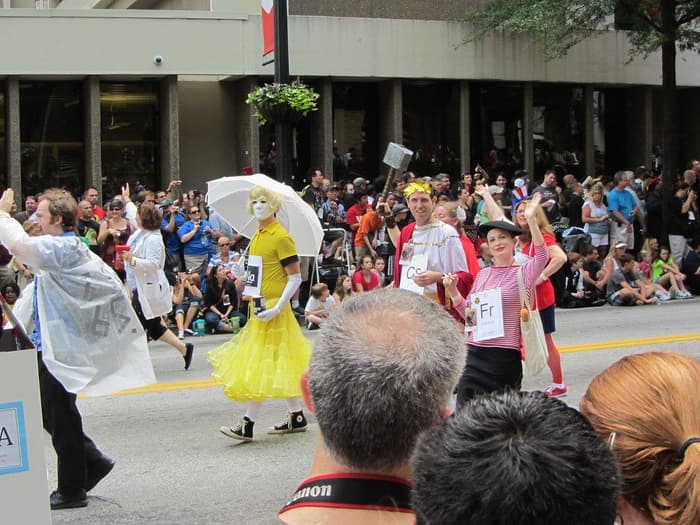 Costumed marchers dressed as elements of the periodic table walking past a packed sidewalk crowd at the Dragon Con parade in downtown Atlanta