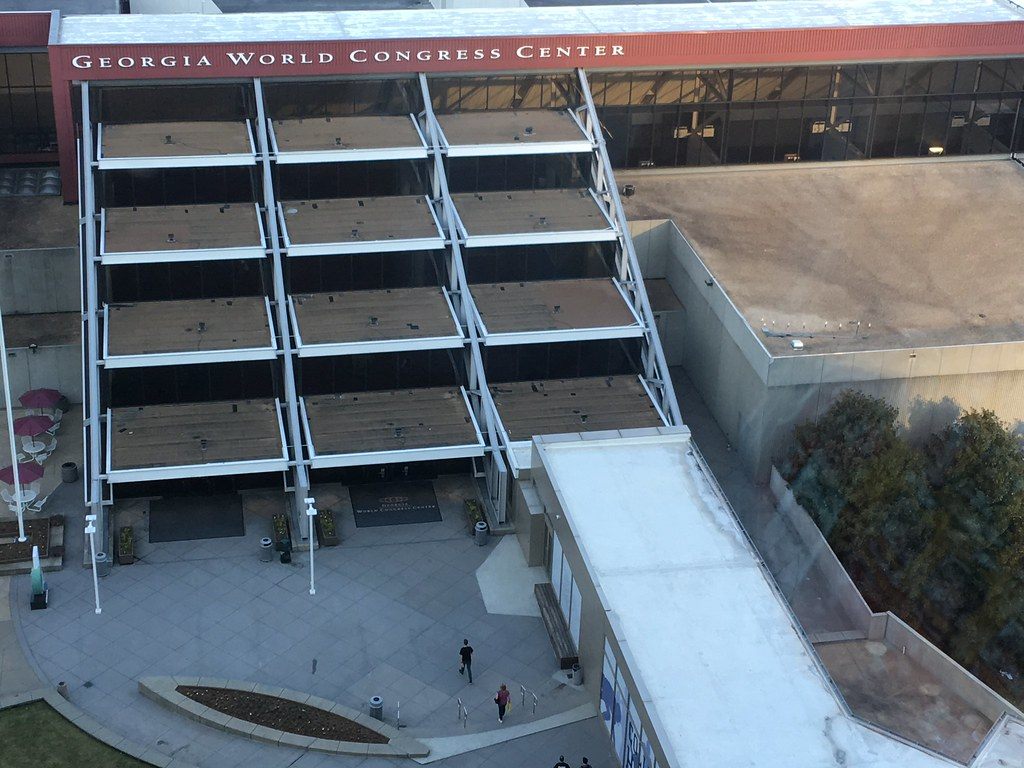 An entrance to the Georgia World Congress Center seen from above, with the center's name across the top of a stepped glass facade and people crossing the plaza