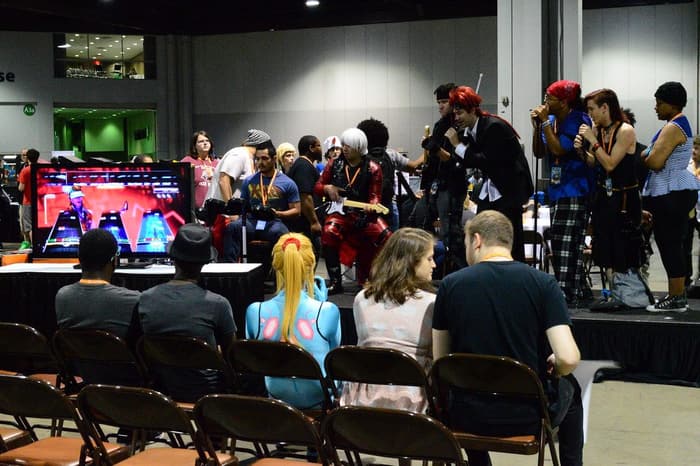 Cosplayers playing a rhythm game on a stage while badge-wearing fans watch from rows of chairs in a Georgia World Congress Center hall during MomoCon 2015