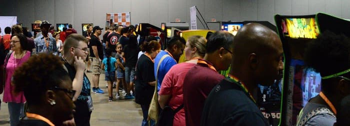 Crowds of MomoCon 2015 attendees playing and queuing along a long aisle of arcade cabinets in a Georgia World Congress Center exhibit hall