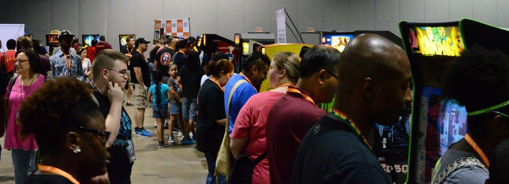 Crowds of MomoCon 2015 attendees playing and queuing along a long aisle of arcade cabinets in a Georgia World Congress Center exhibit hall
