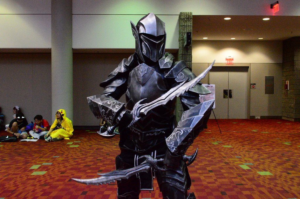 A cosplayer in black Skyrim armor with curved blades in a carpeted GWCC lobby during MomoCon 2015, with fans resting on the floor behind