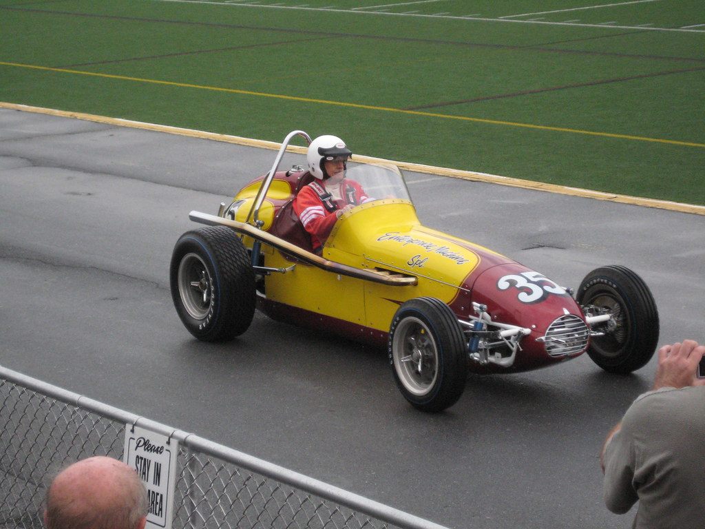 A helmeted driver takes a yellow vintage single-seat race car past spectators at a fence during the 2009 AACA Hershey meet