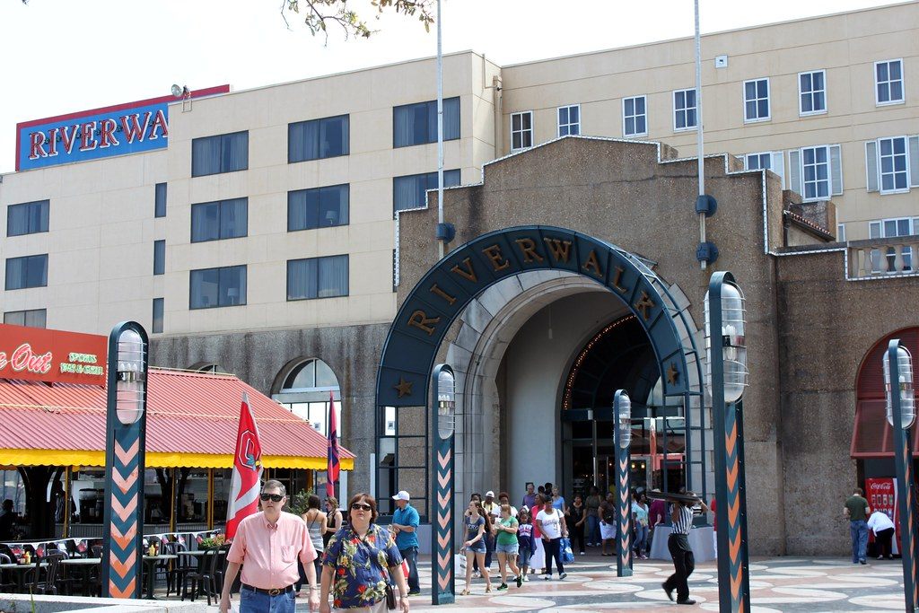 Shoppers walking under the arched Riverwalk entrance of the riverfront mall, with a Riverwalk sign on the building above