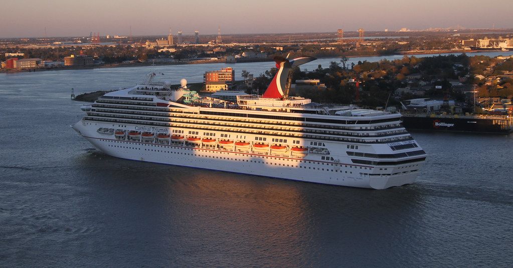 A large cruise ship on the Mississippi River in low evening sun, seen from above, with the far bank behind