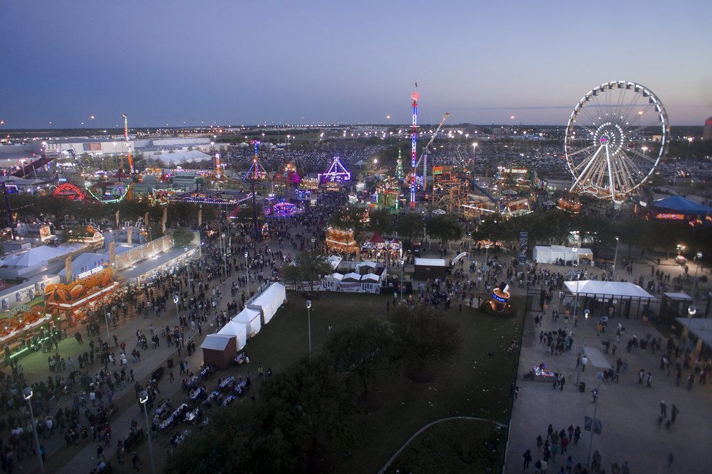 The Houston Livestock Show and Rodeo carnival seen from above at blue hour, crowds between lit rides and a Ferris wheel, with Reliant Park's lots beyond