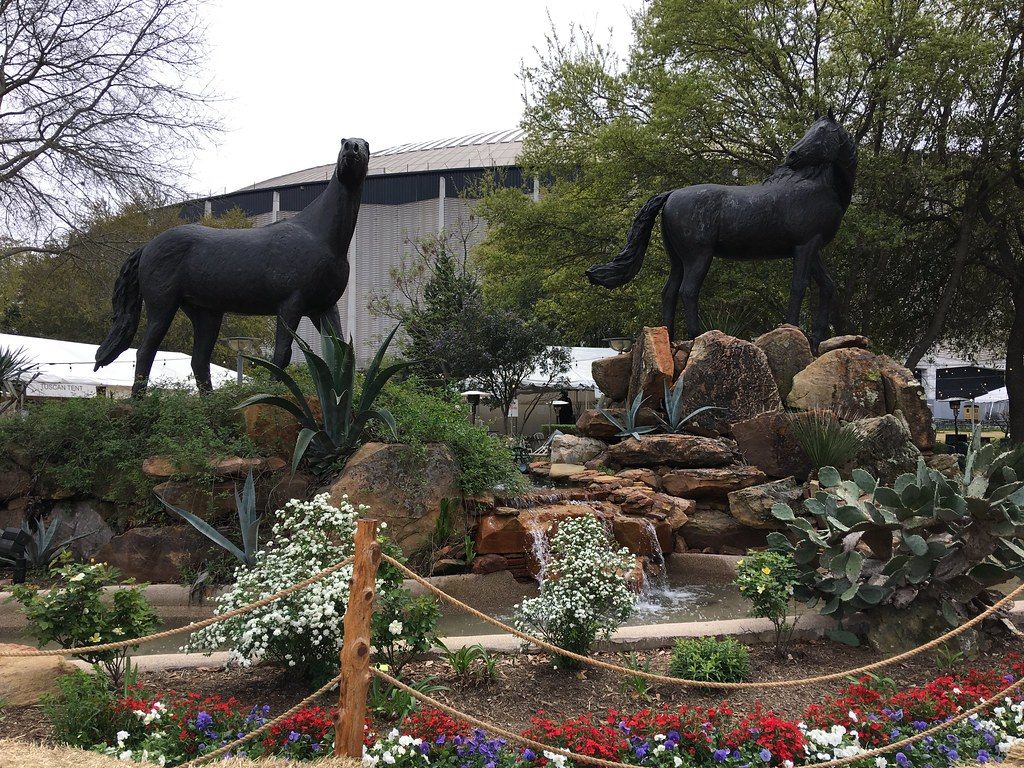Two black horse sculptures on a rock fountain planted with cactus and flowers, with the gray wall of the Astrodome behind the trees