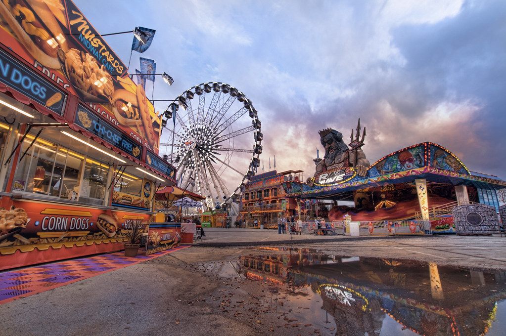Carnival food stands and a Ferris wheel reflected in a puddle under a dramatic evening sky at the Houston Livestock Show and Rodeo carnival
