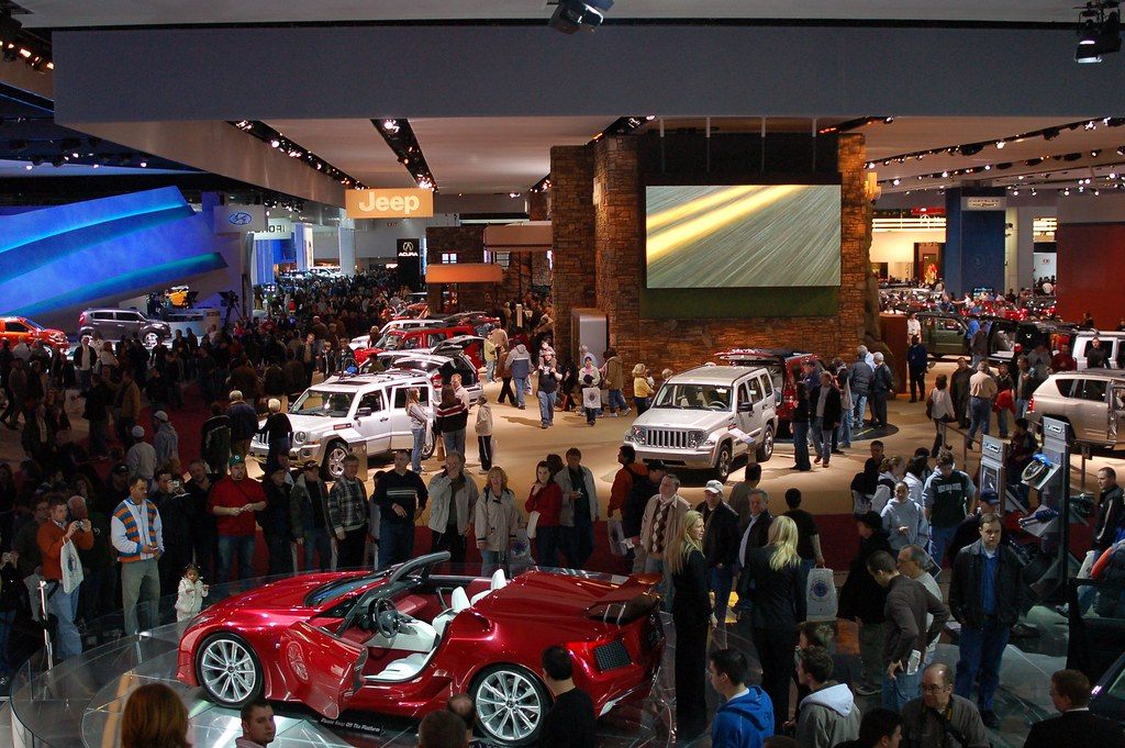 A packed show floor seen from above, with crowds around a red concept roadster, Jeep SUVs and a stone-pillared display at the 2008 North American International Auto Show in Detroit