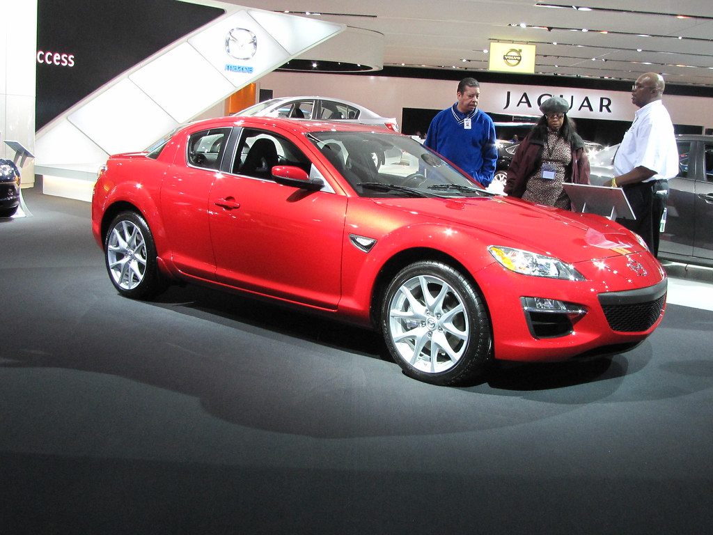 Visitors chatting beside a red Mazda RX-8 on a turntable stand at the North American International Auto Show in Detroit