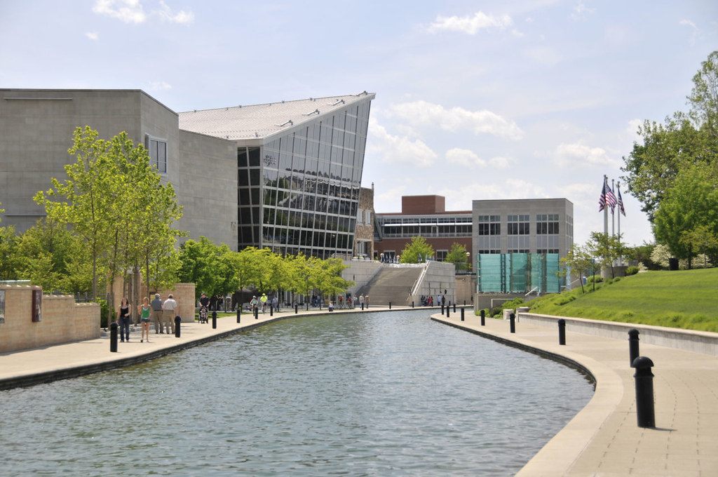 People stroll the paved banks of the downtown Indianapolis Canal Walk past modern museum buildings and young trees