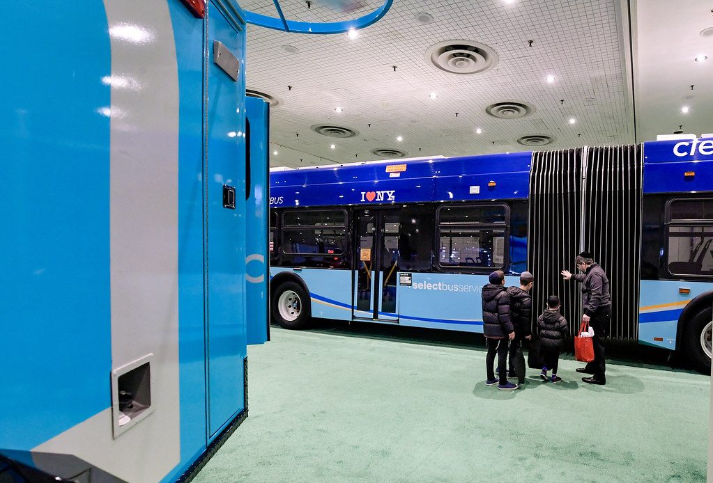 A family looking over an MTA articulated electric Select Bus on the green-carpeted floor of the New York International Auto Show at the Javits Center