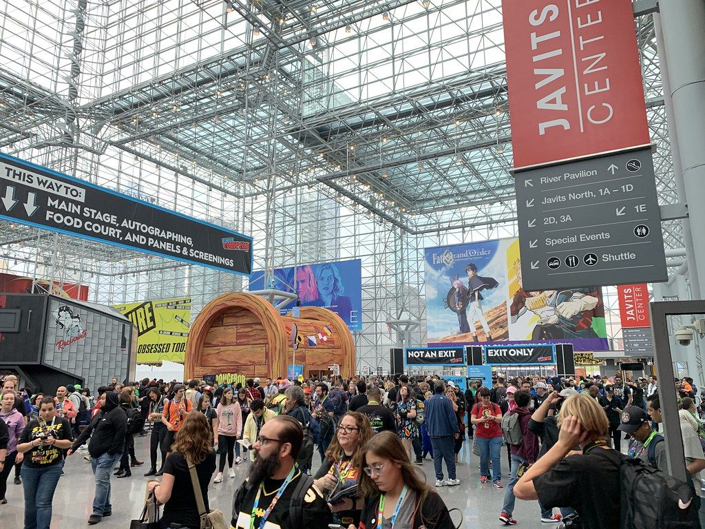 New York Comic Con 2019 crowds under the glass space frame of the Javits Center, beside red Javits Center wayfinding signs for the North halls, River Pavilion and shuttle