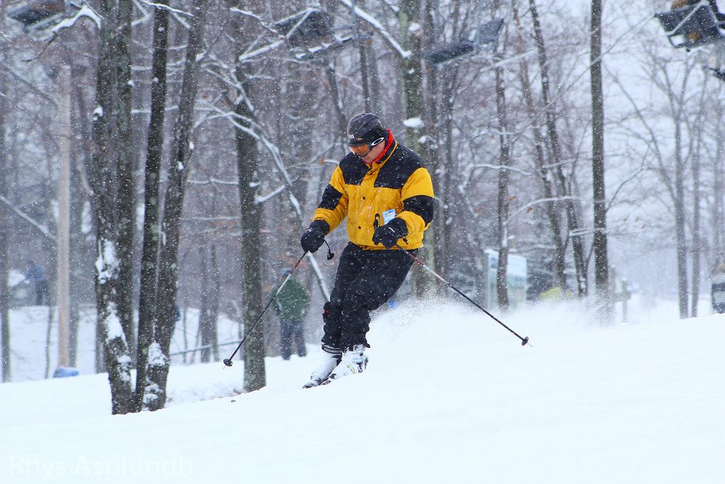 A skier in a yellow and black jacket carving through falling snow between bare trees at Jack Frost Mountain in the Poconos