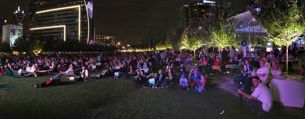 A large crowd on the lawn at Klyde Warren Park at night watching a Dallas Symphony Orchestra simulcast, with lit towers behind