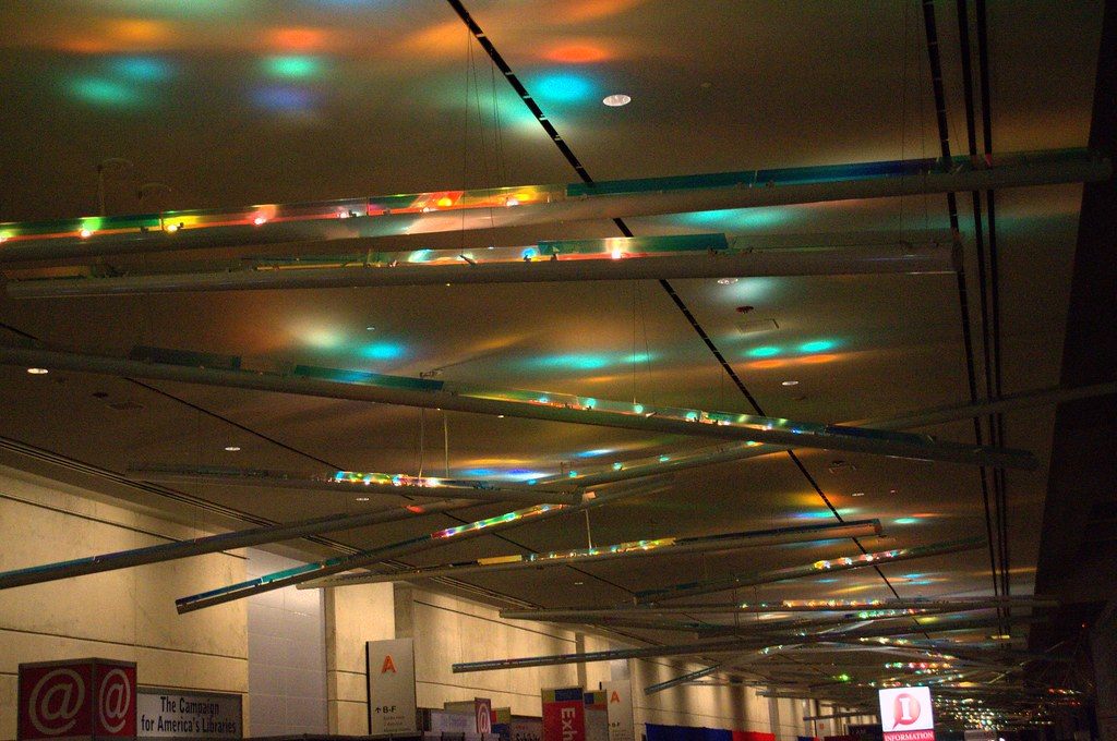 A long convention center hallway with colored light patterns on a low ceiling above meeting-room doors and signs