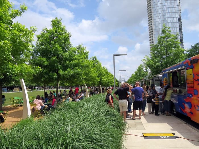 People line up at a brightly painted food truck along a planted walkway at Klyde Warren Park in downtown Dallas