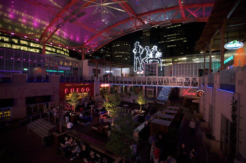 People at tables in the lit courtyard of the Power and Light District at night, under a glass canopy and neon signs