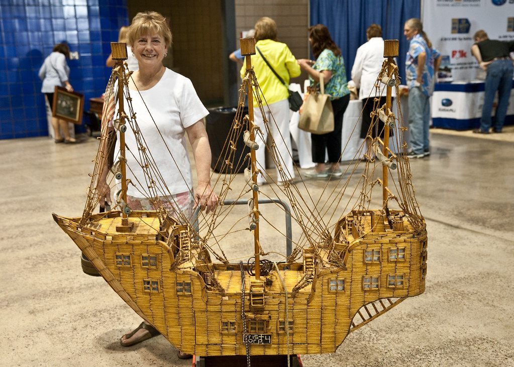 A visitor holding a large model sailing ship in the exhibit hall during the Antiques Roadshow taping at Bartle Hall, with other guests and blue drape behind