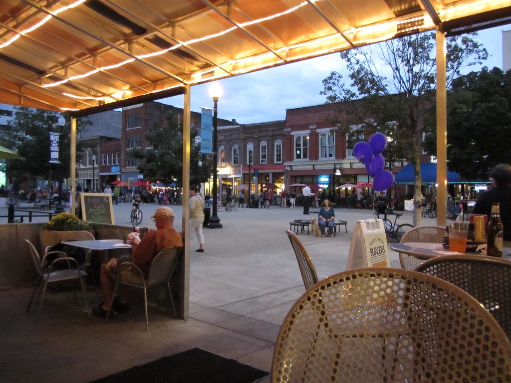 Diners on a covered patio look out over Market Square in downtown Knoxville at dusk, with crowds among the brick storefronts