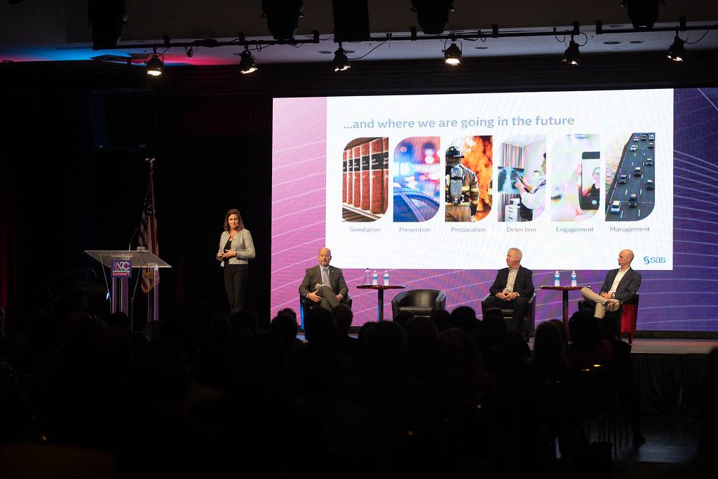 A packed, darkened room watches a four-person panel on stage under a big screen at a general session of the 2024 N.C. Transportation Summit at the Koury Convention Center