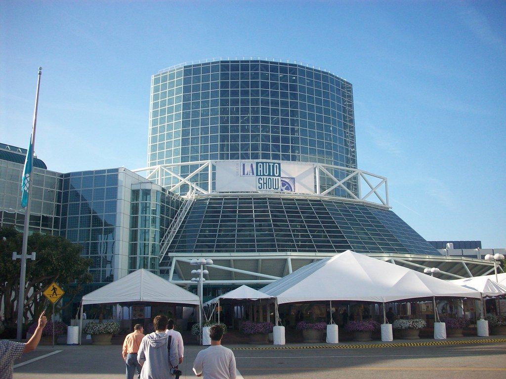An LA Auto Show banner on the LACC's glass tower, with white tents on the plaza in front