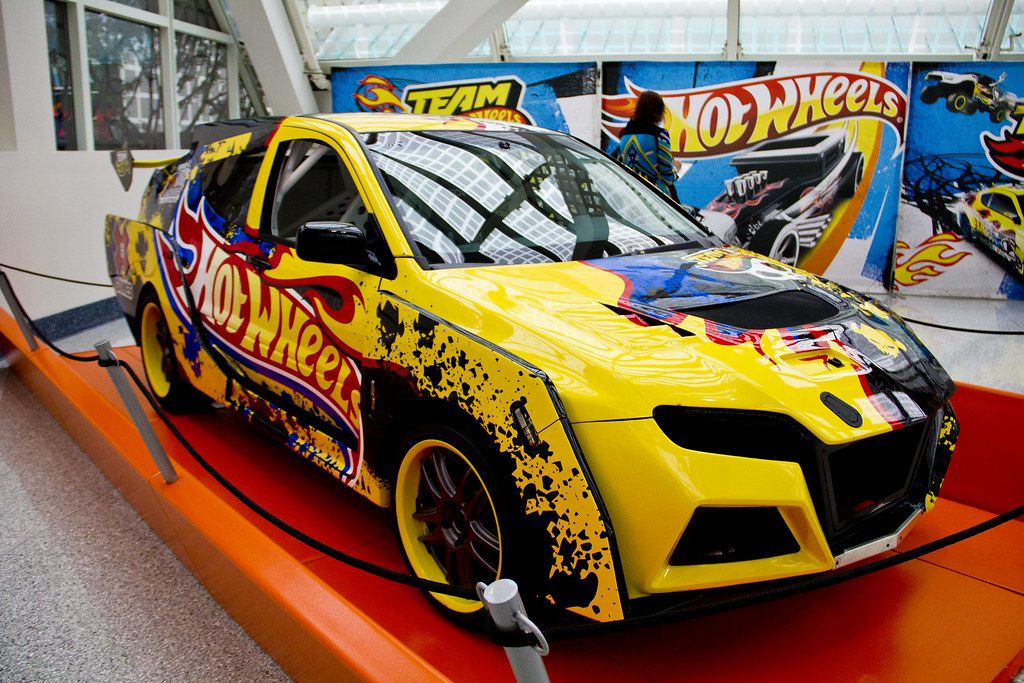 A yellow Hot Wheels-wrapped concept car on an orange display under the glass lobby at the 2012 LA Auto Show