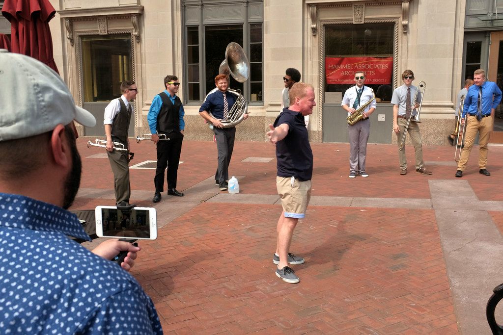 A man dancing on the brick plaza of Penn Square in Lancaster while a brass band plays and a friend films on a phone