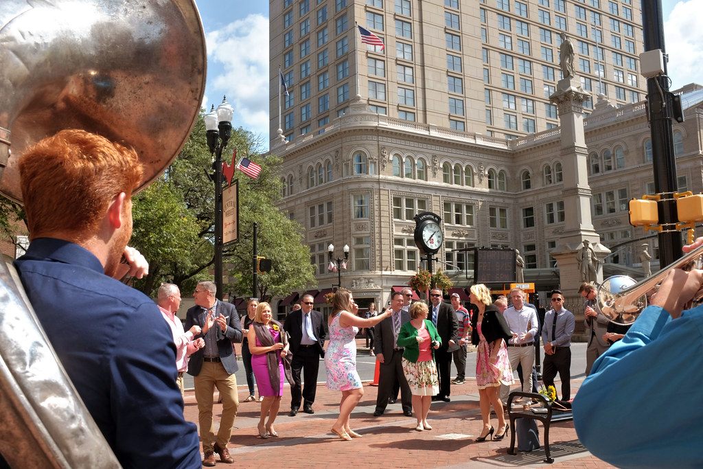 A sousaphone player leads a dancing crowd across Penn Square in Lancaster, with a street clock and a high-rise tower behind