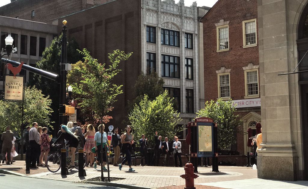 A dressed-up crowd walking along the brick sidewalk of Penn Square in downtown Lancaster beside a Central Market sign