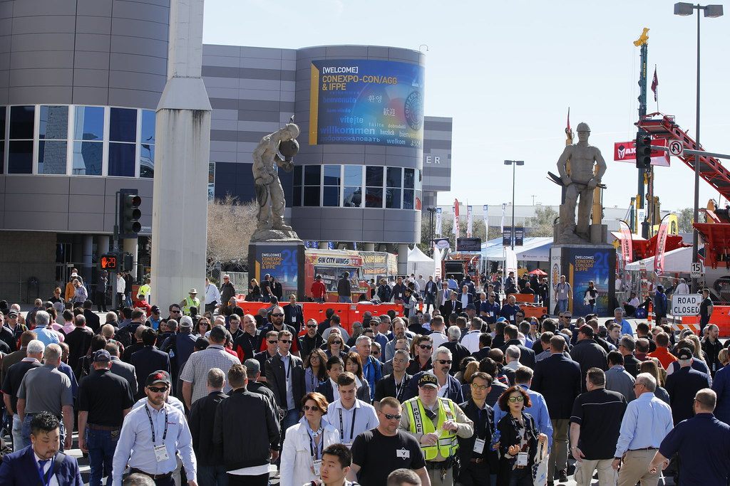 A dense crowd of badge-wearing attendees walking out of the Las Vegas Convention Center plaza under a CONEXPO-CON/AGG and IFPE welcome screen, with equipment displays and cranes behind