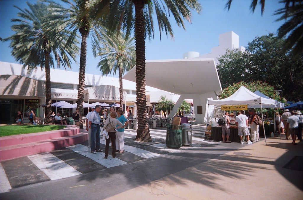 Shoppers stroll under palm trees past white market tents and a 1960s shade canopy on Lincoln Road Mall