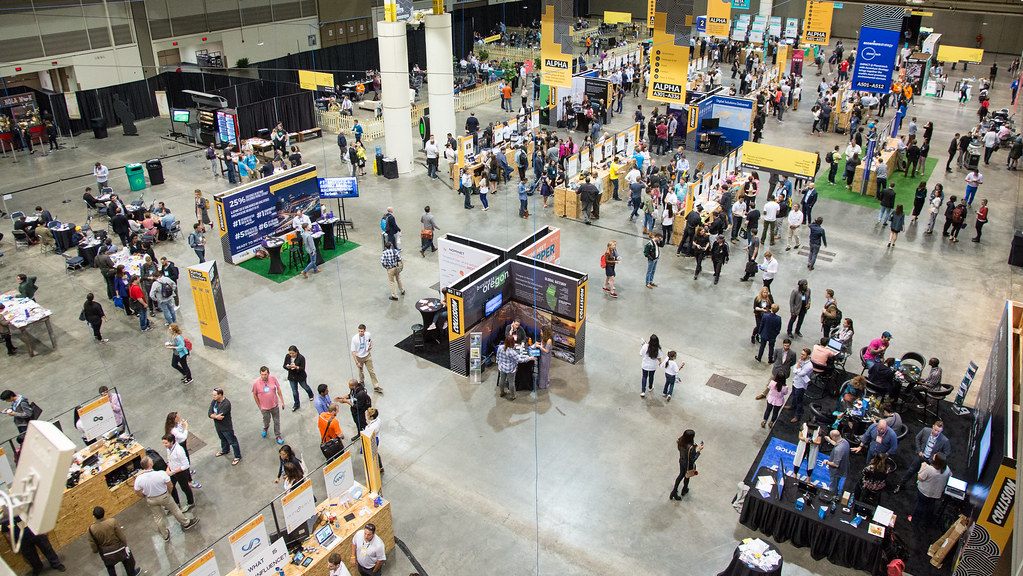 Looking down on a busy exhibit hall with hundreds of attendees, startup booths and yellow banners at the Collision conference in New Orleans