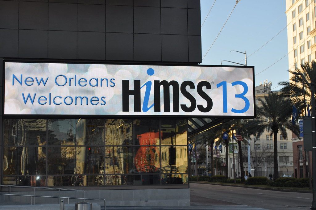 A New Orleans Welcomes HIMSS13 sign over the dark glass of the convention center, with palm trees along the street