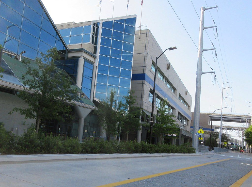 The blue glass and pale panels of a Morial Convention Center wing above young trees along a wide sidewalk