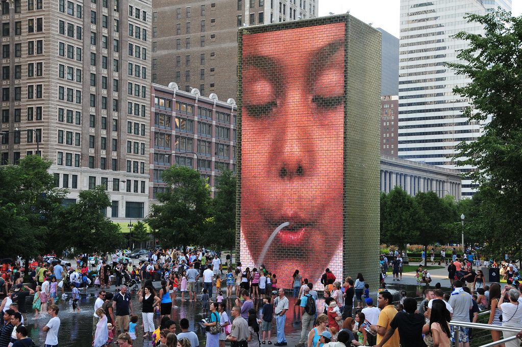 A summer crowd wading and splashing at the Crown Fountain in Millennium Park as a giant video face spouts water, with Michigan Avenue towers behind