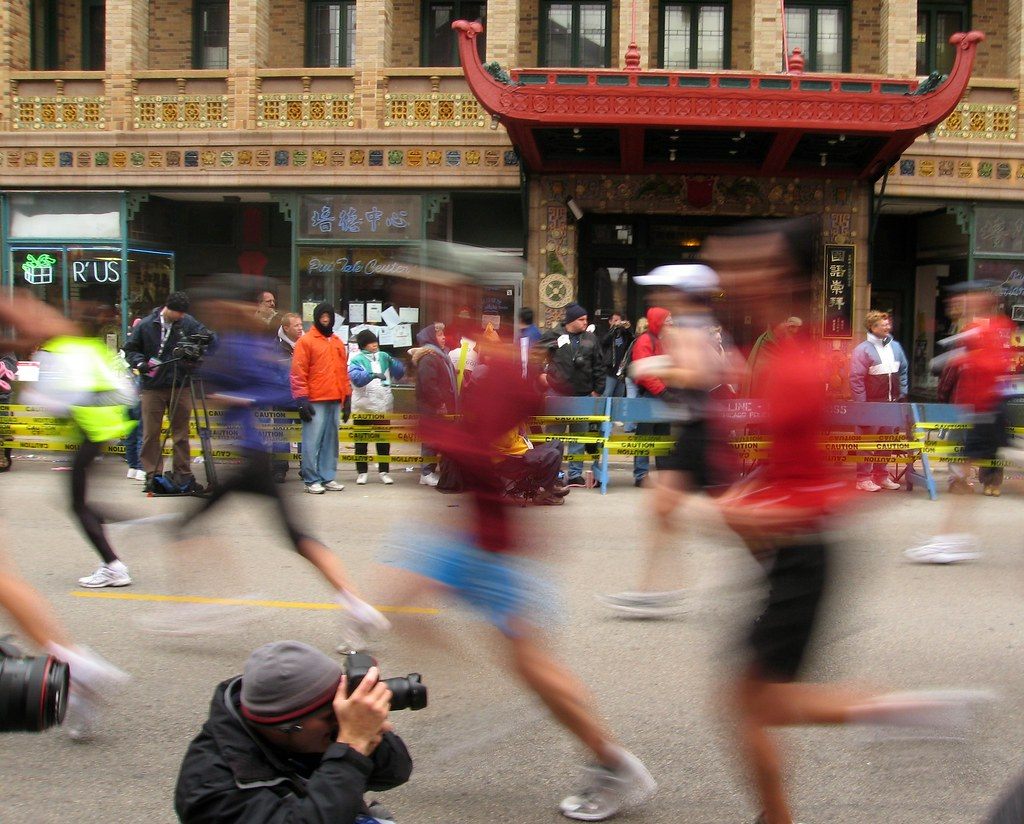 Chicago Marathon runners blurring past spectators and a photographer under the red pagoda-style canopy of a building in Chinatown