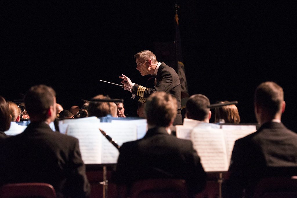 A U.S. Navy Band conductor in dress uniform leads musicians on a darkened stage during a March 2016 concert at the Mid-Hudson Civic Center