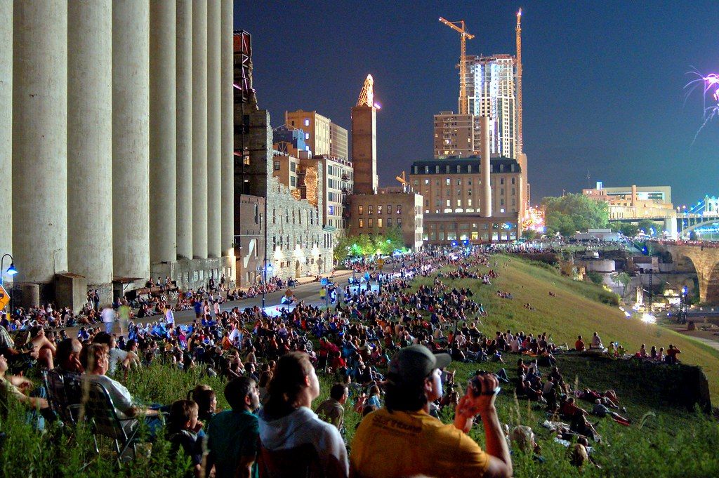 A big crowd packed on a grassy riverbank at dusk below the downtown Minneapolis skyline, waiting for Aquatennial fireworks