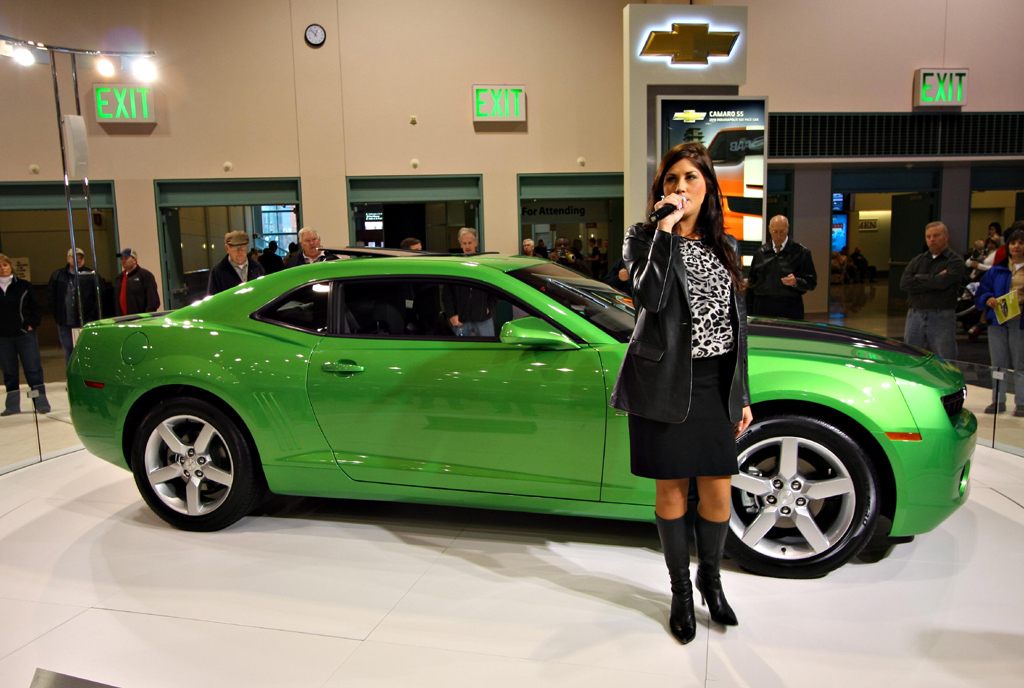 A presenter with a microphone beside a green Chevrolet Camaro as visitors watch at the 2010 Twin Cities Auto Show