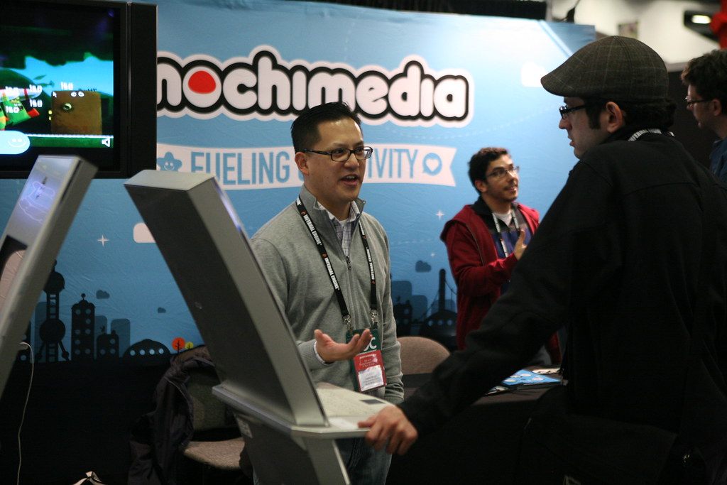 An exhibitor talking with badge-wearing visitors at a game-publisher booth with kiosks and screens during GDC 2011 at Moscone Center