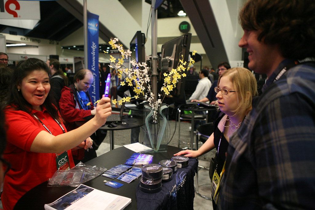 A smiling exhibitor in a red GDC shirt demonstrating a gadget to laughing visitors across a booth table on the GDC 2011 expo floor at Moscone Center