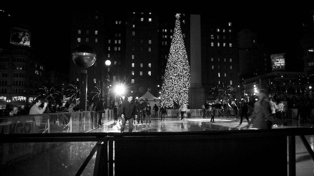 Black and white night view of skaters on the holiday ice rink in Union Square, San Francisco, with a lit Christmas tree and hotel towers behind