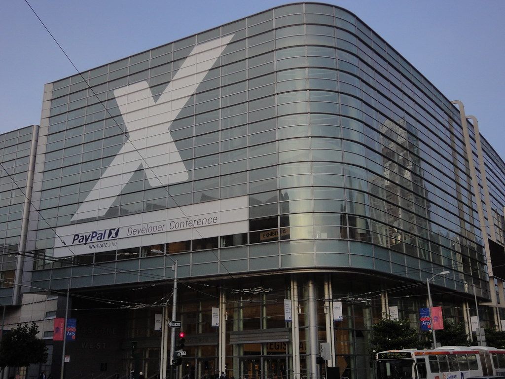 The curved glass facade of Moscone West wrapped in a giant X graphic and a PayPal X Developer Conference banner, with a Muni bus passing below