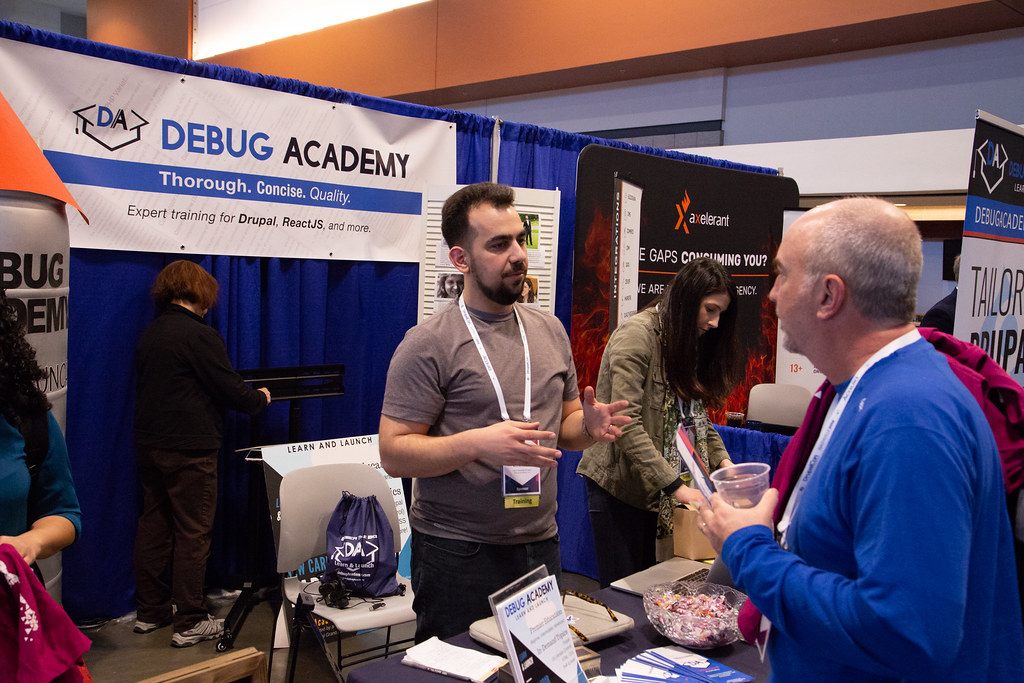 An exhibitor talks with a visitor at the Debug Academy booth, DrupalCon Nashville 2018