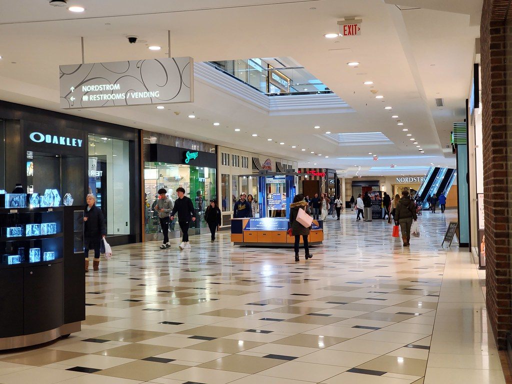 Shoppers walking a wide tiled concourse past an Oakley store toward Nordstrom inside Twelve Oaks Mall, Novi