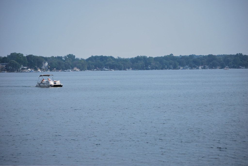 A pontoon boat crossing Walled Lake on a hazy summer day, with lakeside houses and trees on the far shore