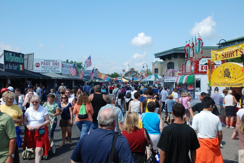 Crowds walking down a fairgrounds street lined with food stands, flags and souvenir booths during the New York State Fair