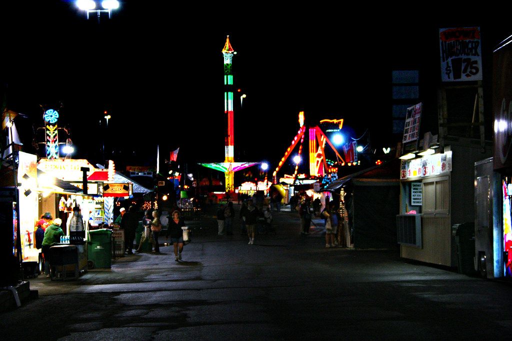 The fairgrounds midway at night, with lit rides and a drop tower at the end of a row of food stands